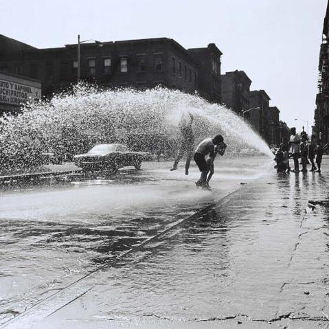 A photograph of kids playing in the street with a fire hydrant splashing water.