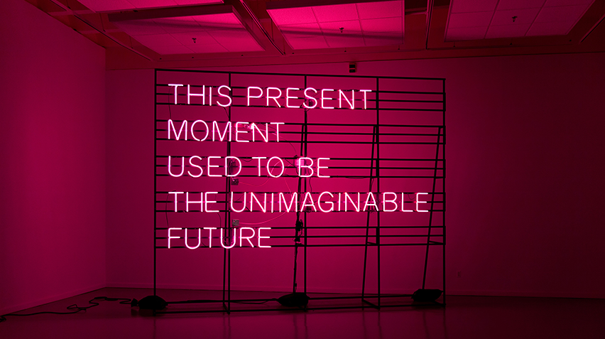 A pink neon sign that reads "This Present Moment Used to be the Unimaginable Future."