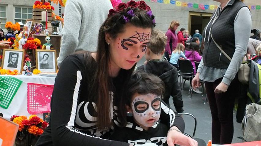 A mother and child, whose faces are painted for the Day of the Dead, sit at a table and participate in crafts