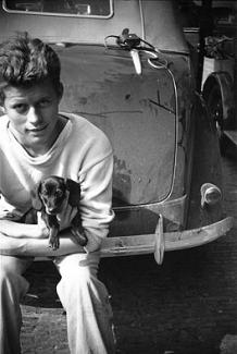 A young Jack Kennedy sitting on the bumper of a car, holding a puppy