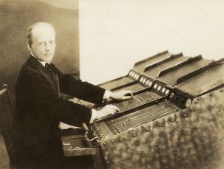 A sepia toned photograph of Thomas Wilfred sitting at a machine called a Clavilux, which he toured the country with creating art. 