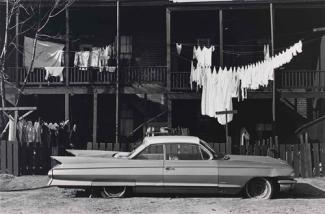 A gelatin silver print of a street with a car in the foreground, clothes handing in the middle ground and a building in the background.