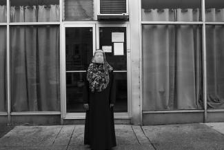 Black and white photo of an African American woman wearing a swirl-printed hijab and protective face shield stands outside a buidling with large plate-glass windows.