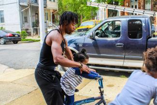 A muscular African American man helps a small child ride a large bike. Another child can be seen at the extreme foreground. Work trucks are parked along the street.