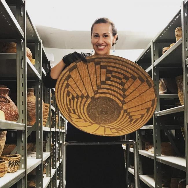 A photograph of a woman holding a large basket in museum collection storage.