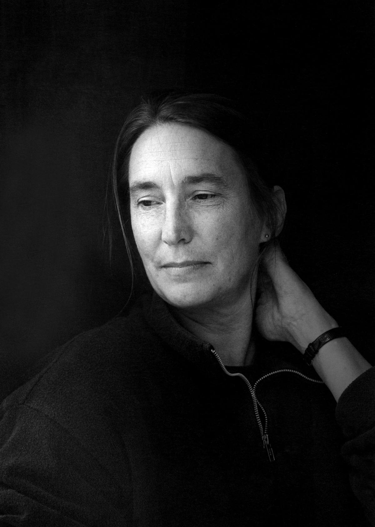 Bio Image - Jenny Holzer Black and white photo of Jenny Holzer with a light source to the left and her left hand on the back of her neck.