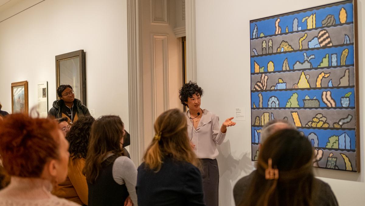 Press - Research and Scholars "Art Bites" Program A person stands in front of an abstract painting. She is speaking to a group of onlookers.