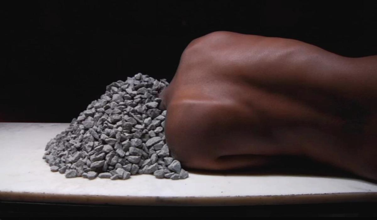 A still from the video shows a woman reclining with her head covered in stones