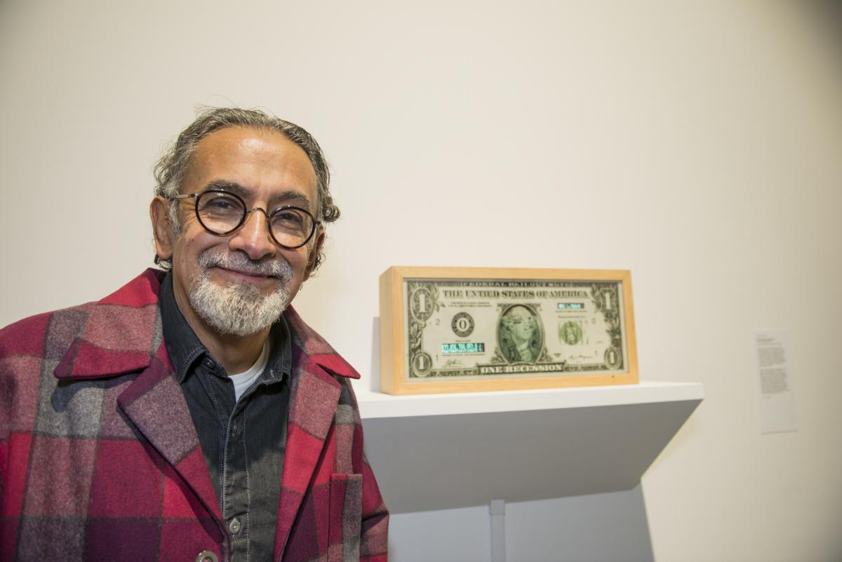 A man with a white beard standing in front of a framed one dollar bill