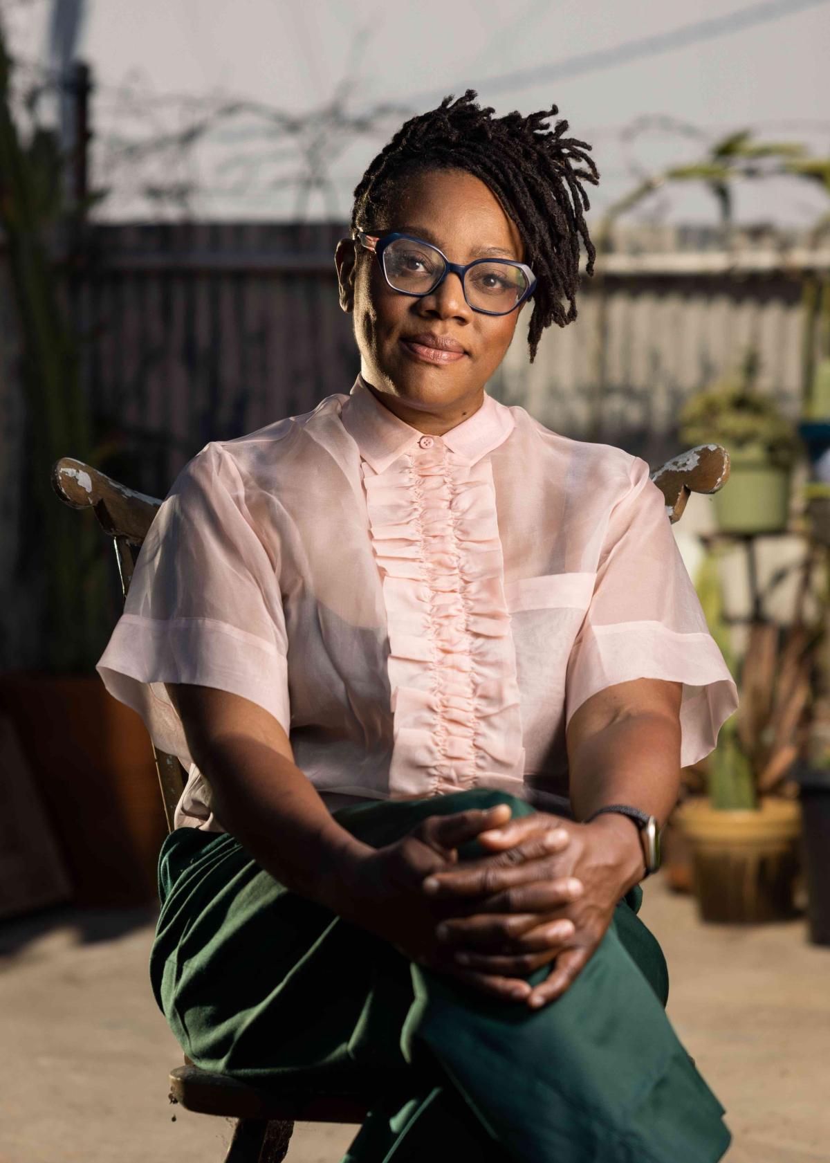 The artist Cauleen Smith sits in a chair in a pink top and green pants