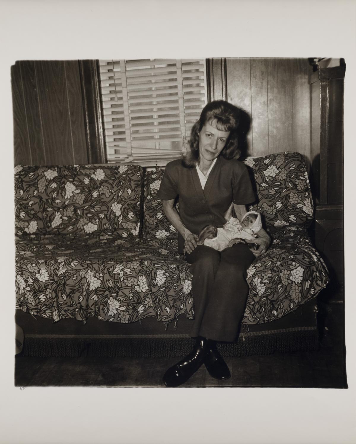 A black and white photograph by Diane Arbus titled "Mrs. Gladys 'Mitzi' Ulrich with the baby, Sam, a stump-tailed macaque monkey"