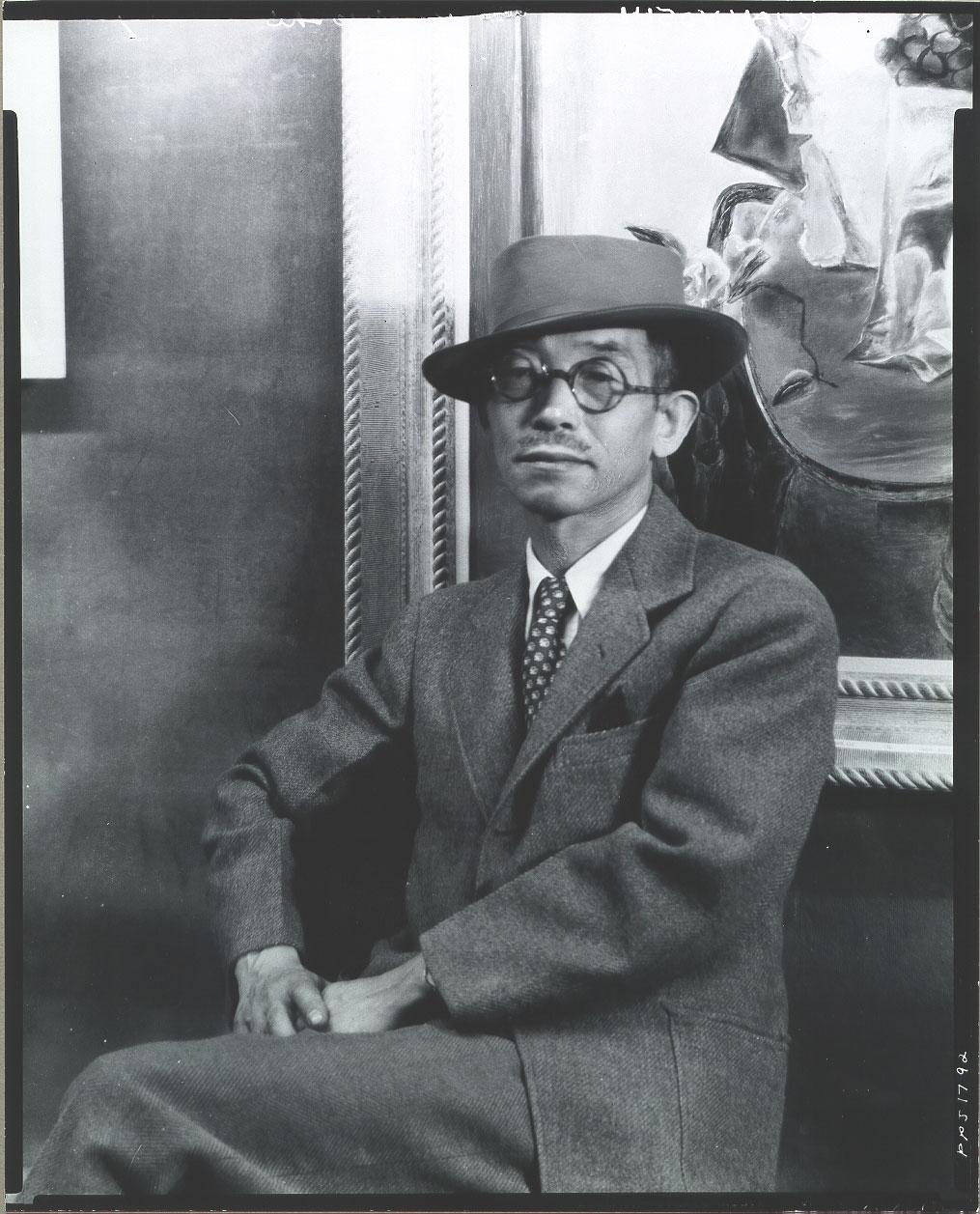 A picture of Yasuo Kuniyoshi in a suit and hat sitting in front of artwork.