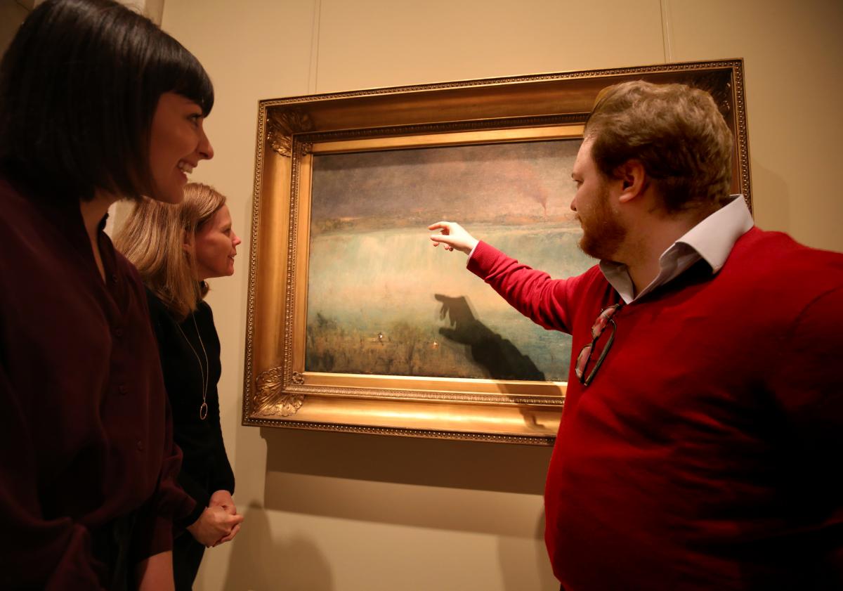 A photograph of a man in a red sweater pointing to a painting of Niagra Falls as others watch.