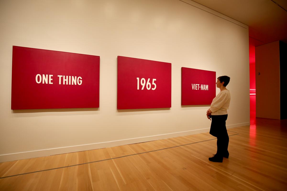 A photograph of a woman standing in front of a three piece painting that says "One Thing, 1965, Viet Nam"