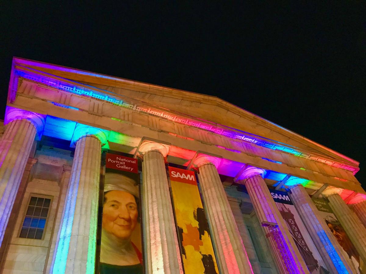 A photograph of the outside of the Smithsonian American Art Museum at night with rainbow lights shining on the building. 
