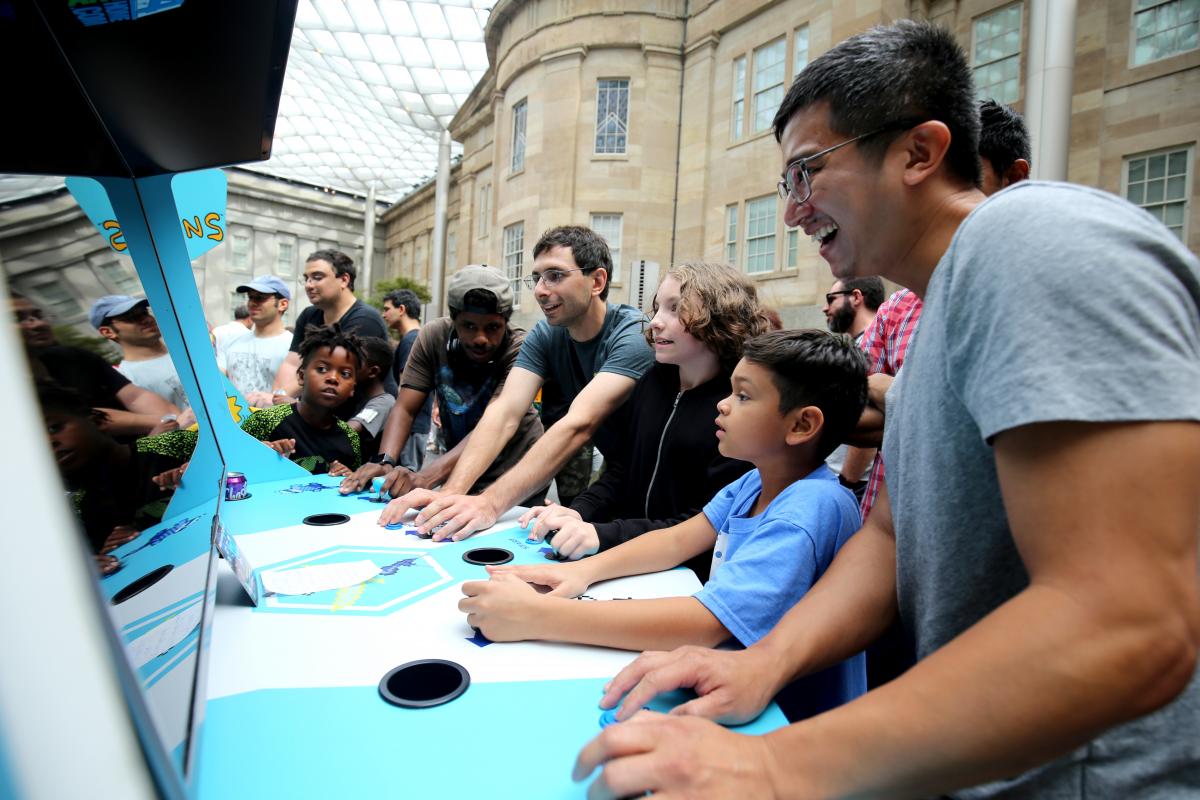 A photograph of six people playing arcade games. 