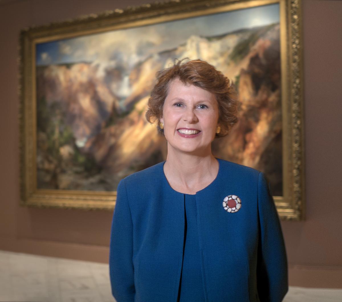 A photograph of a woman standing in blue in an art gallery with a large painting behind her. 