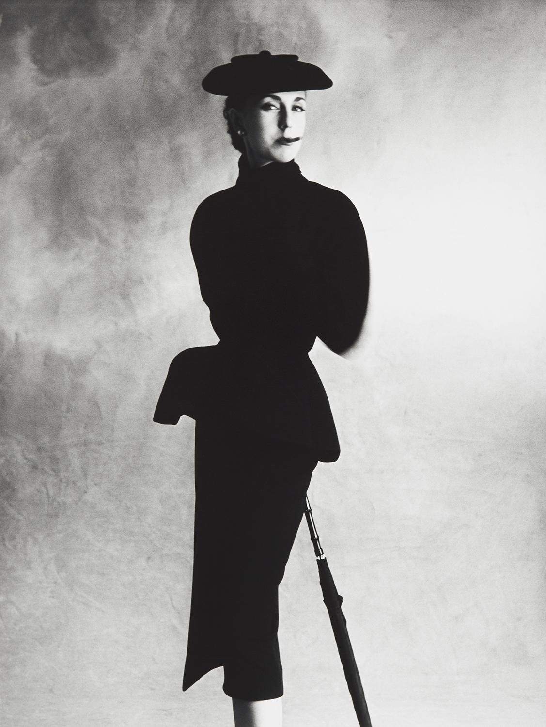 A woman standing in a black suit with a black hat. 