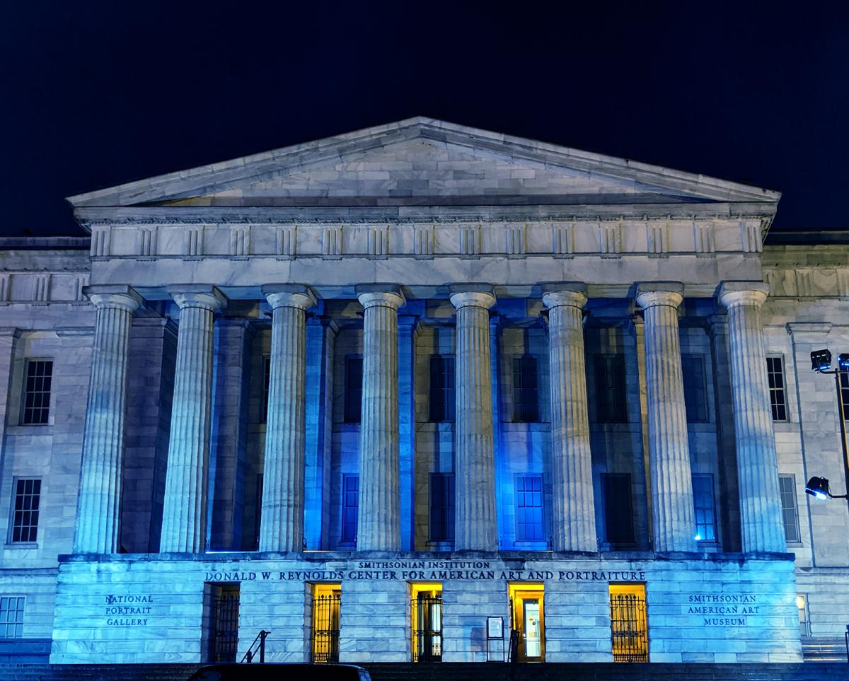 A photograph of the facade of the Smithsonian American Art Museum at night with blue lights honoring healthcare workers.