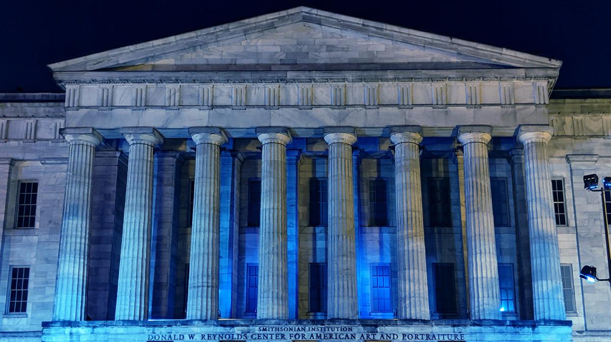 A photograph of the facade of the Smithsonian American Art Museum at night with blue lights honoring healthcare workers.