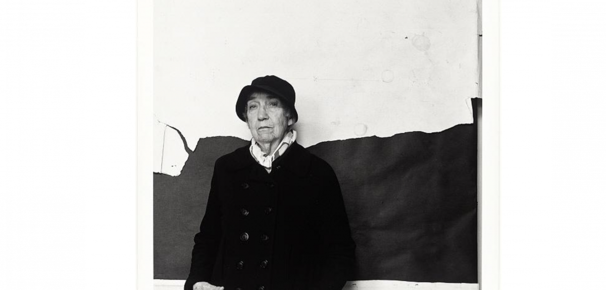 A black and white portait of a woman in a hat and coat standing against a wall.