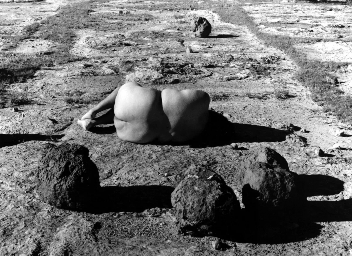 A black and white photograph showing the back of a nude woman lying down on the ground, surrounded by rocks.