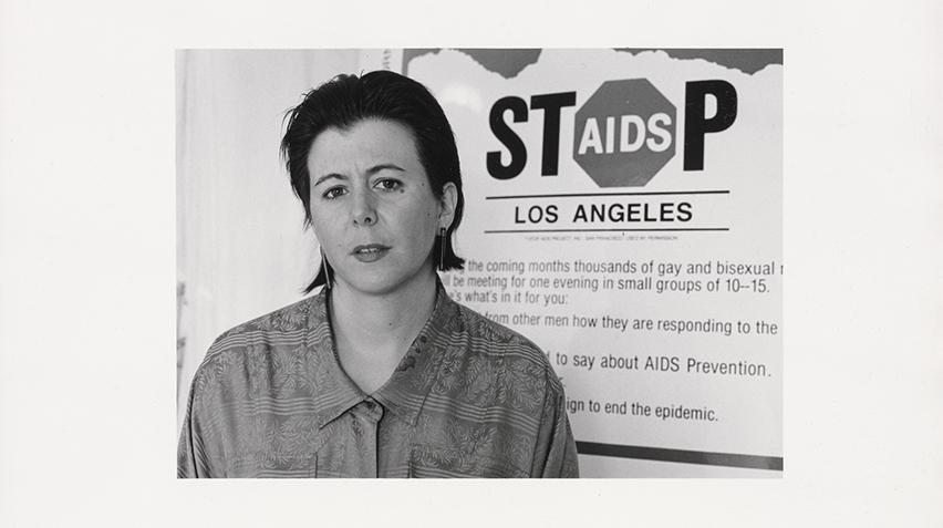 A black and white photograph of a person with short hair standing in front of a sign that says "Stop AIDS Los Angeles."