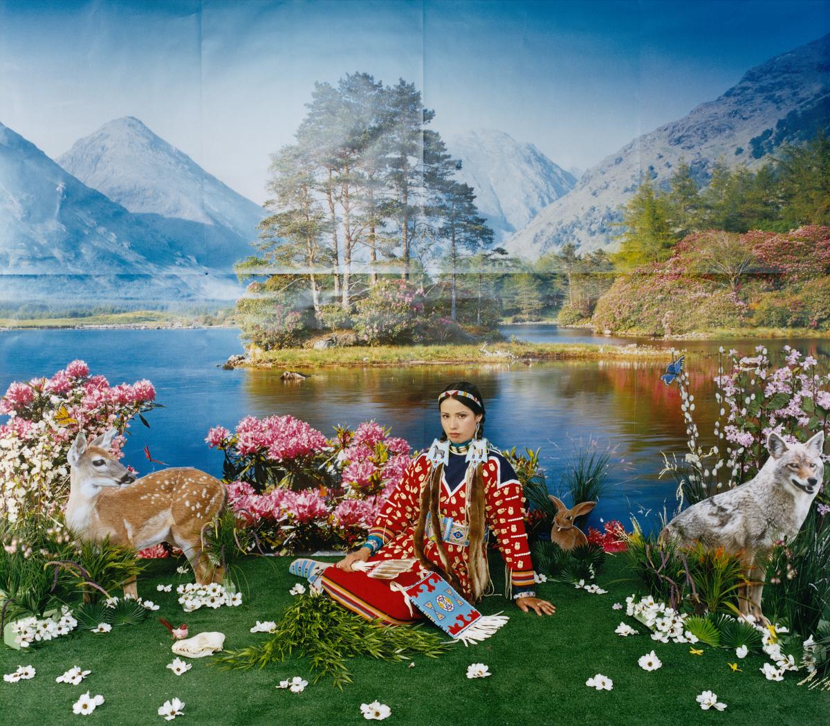 Native woman sitting on a grassy patch with a blow up deer next to her and a mountain landscape