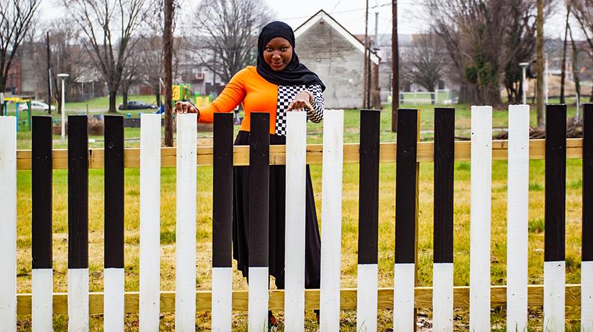 An African American girl wearing a black hijab looks directly at the camera, smiling. In front of her is a fence painted to resenble piano keys. Her fingers rest atop the fence.