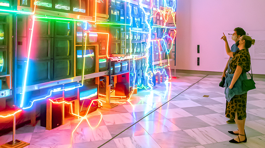 Blog - Art Moments Super Highway, homepage crop Two visitors wearing cloth masks stand in front of the "Electronic Superhighway" installation. One is pointing at the artwork.