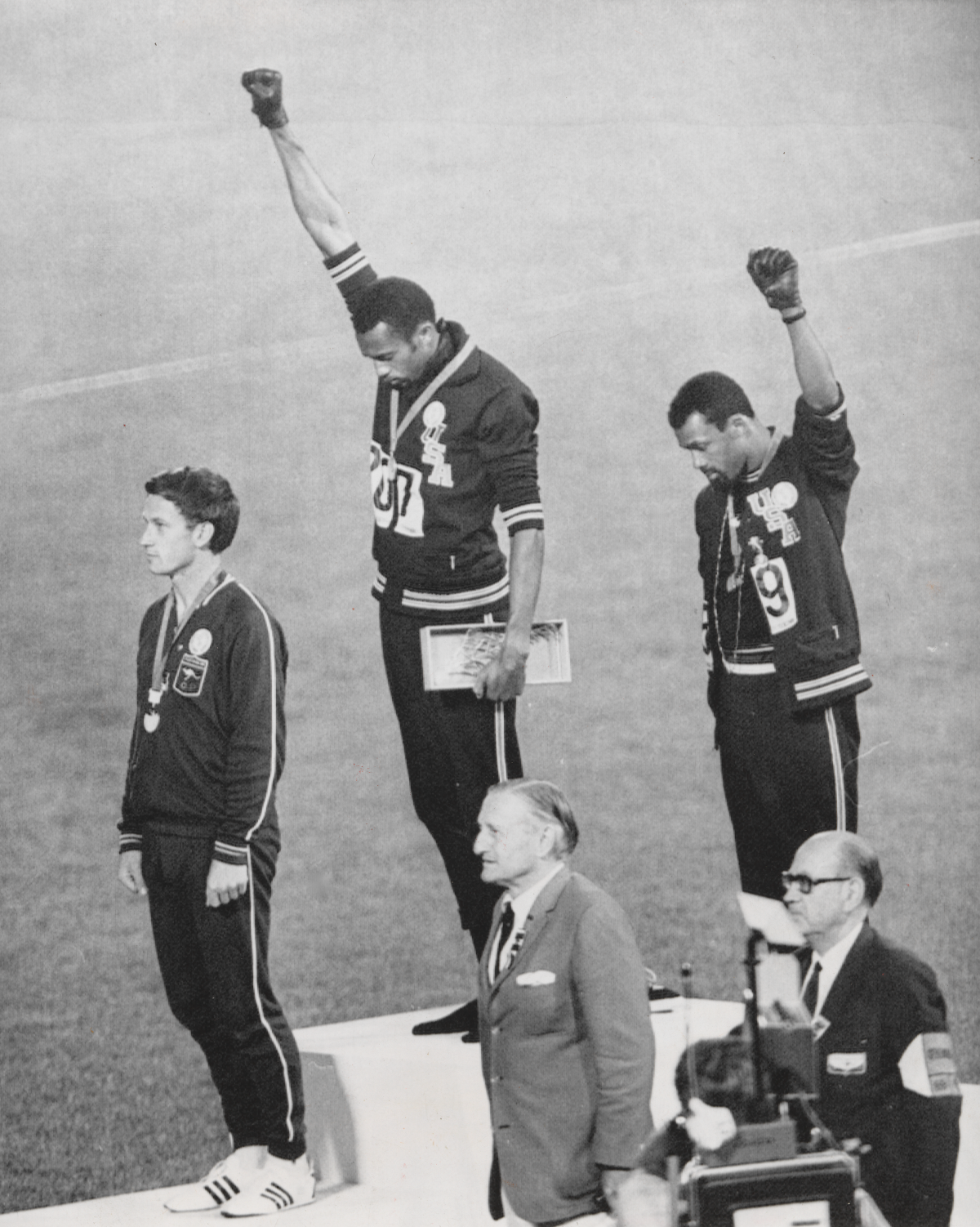 Black and white photograph of Olympic athletes on the podium. Two are holding their fists up in salute.