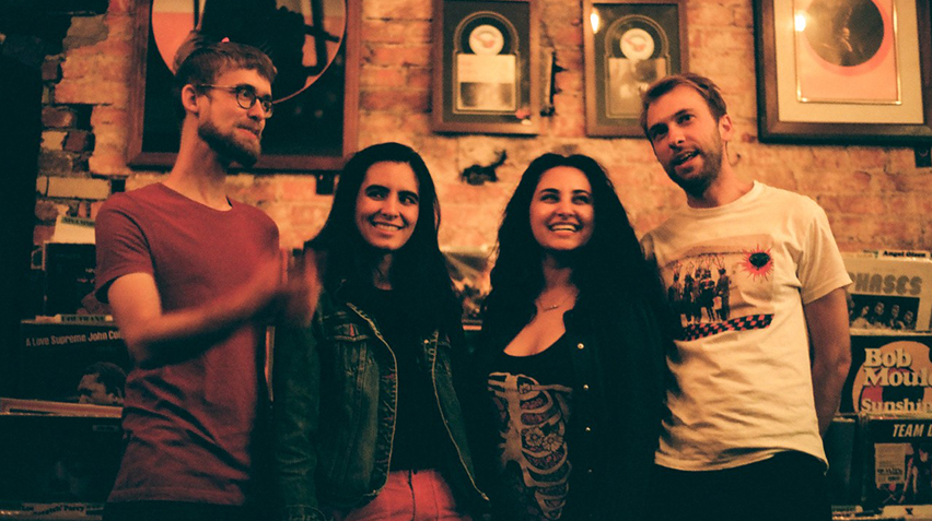 The four musicians of the band Rosie Cima and What She Dreamed stand with their arms around eachother in a room full of framed pictures.