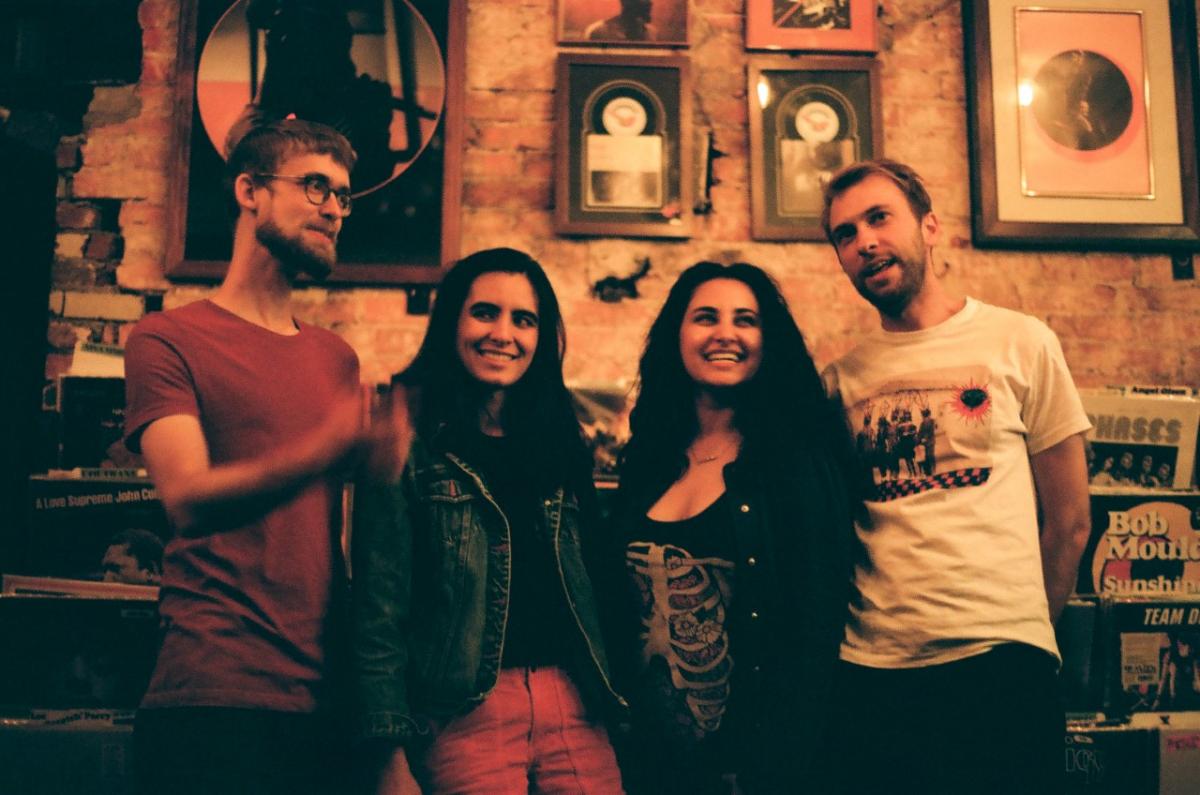 The four musicians of the band Rosie Cima and What She Dreamed stand with their arms around eachother in a room full of framed pictures.