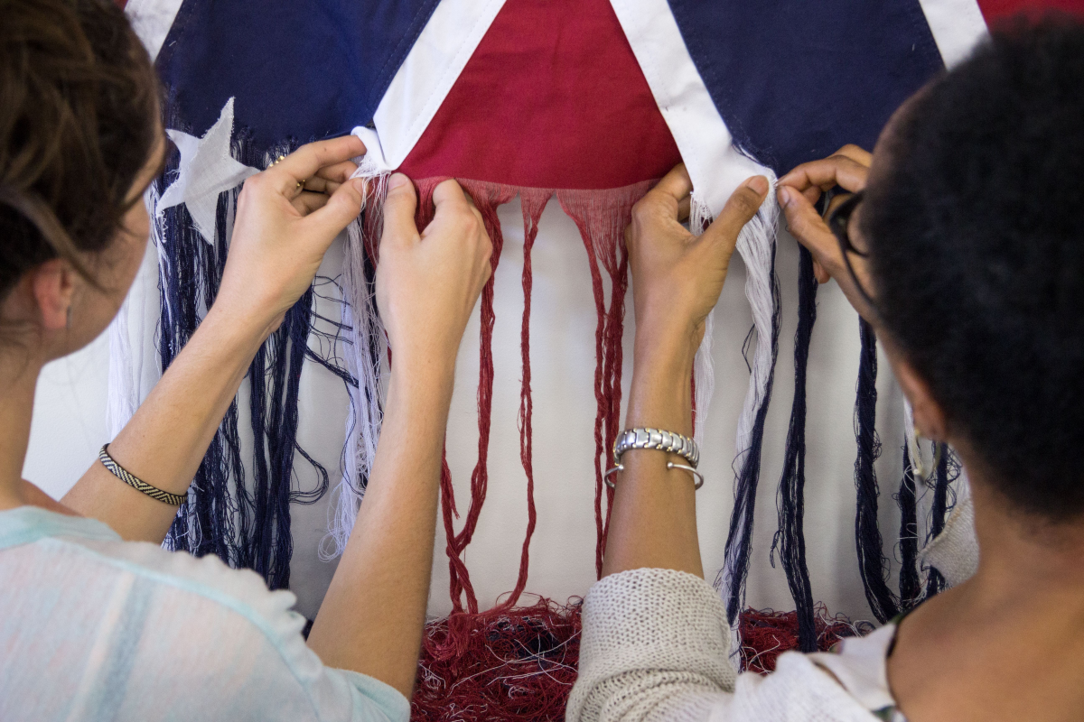 A close up of two pairs of hands on fabric. They are unraveling a Confederate flag.