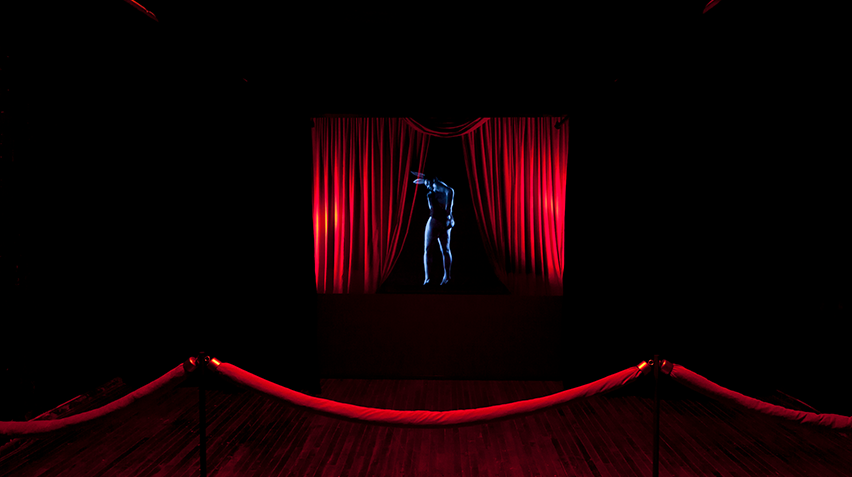 A holographic figure in a dark room behind a velvet rope and drapes.