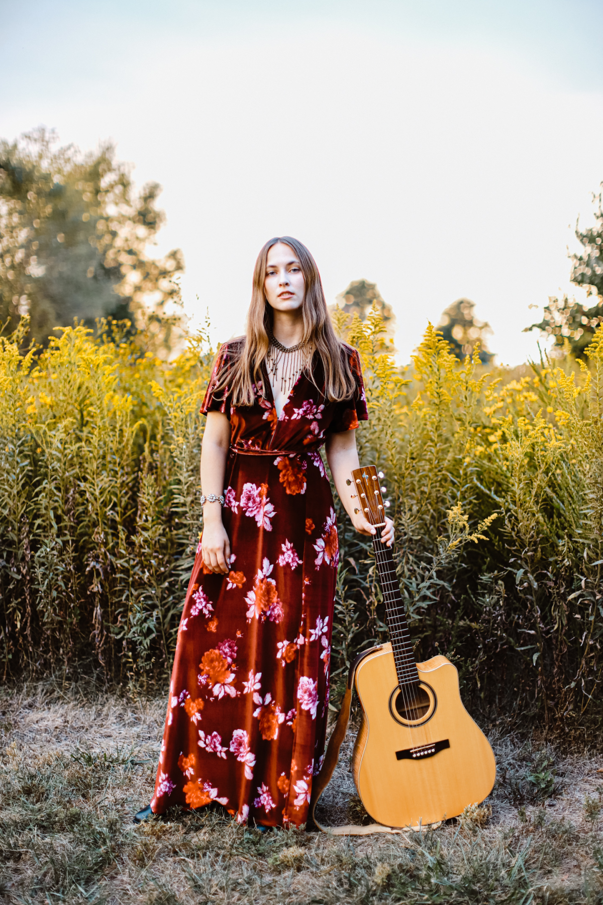 Maddi Mae stands in front of a field of grass wearing a red dress and holding a guitar.
