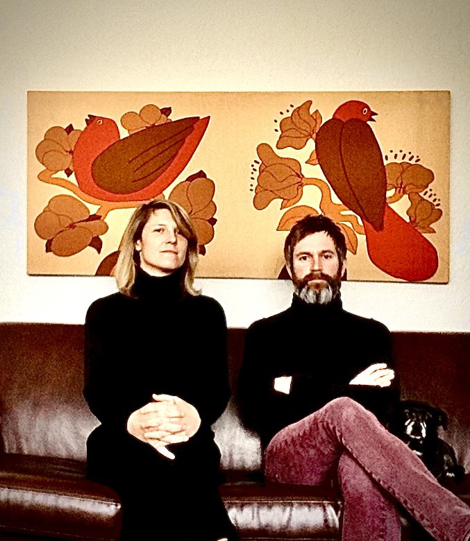 The two members of Astra Via sit on a couch wering matching black turtleneck sweaters.