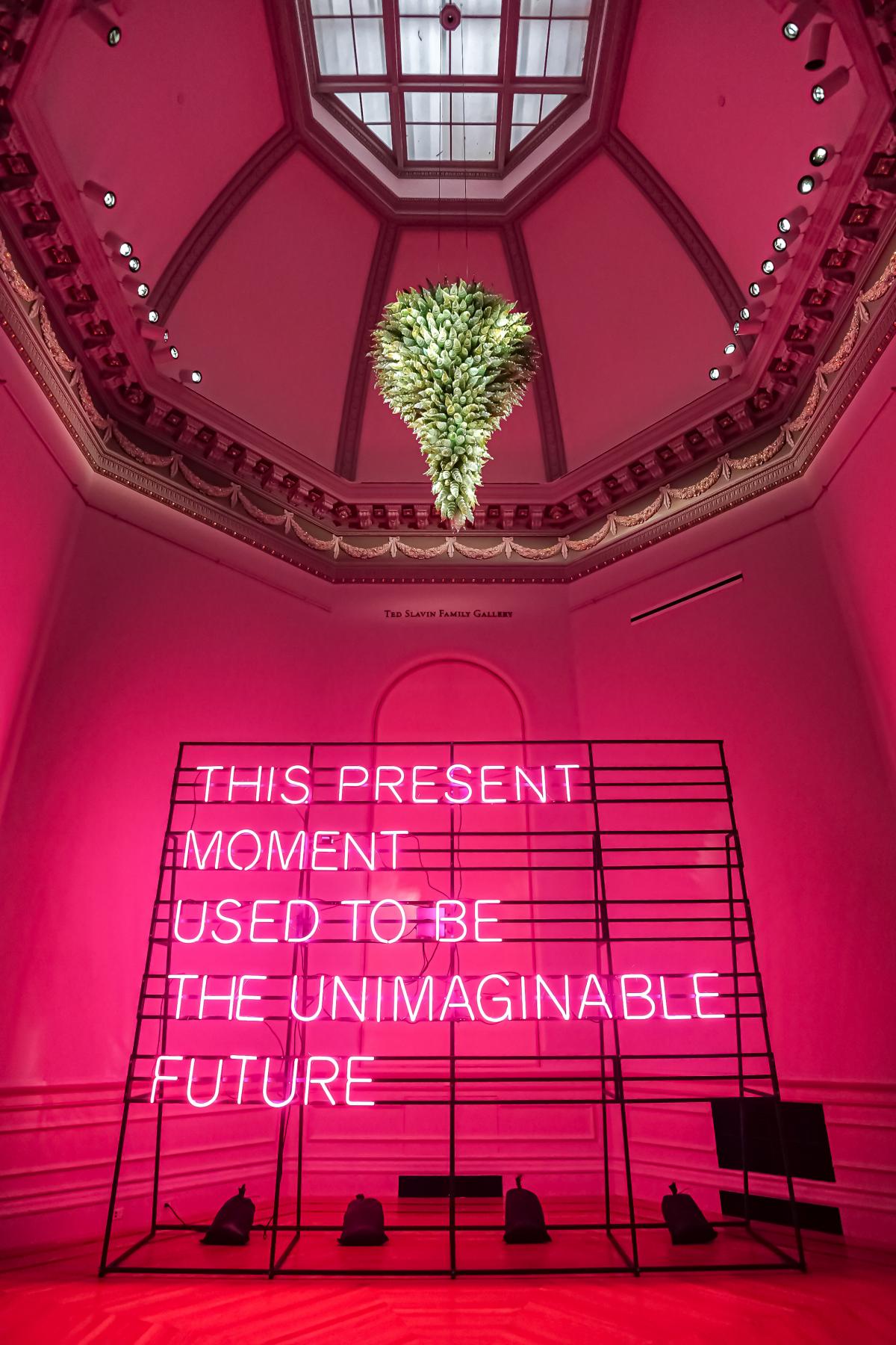 Neon that says "This Present Moment Used to Be the Unimaginable Future" under Chihuly chandelier