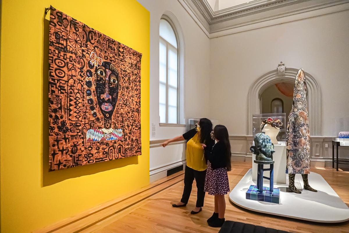 An adult and child standing on a quilted portrait that hangs on a bright yellow wall