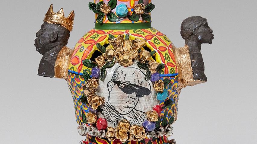 A detailed crop of a highly textured ceramic vessel. The center shows a painting of the rapper Notorious B.I.G. The side handles are busts of Notorious B.I.G. on one and Tupac Shakur on the other.