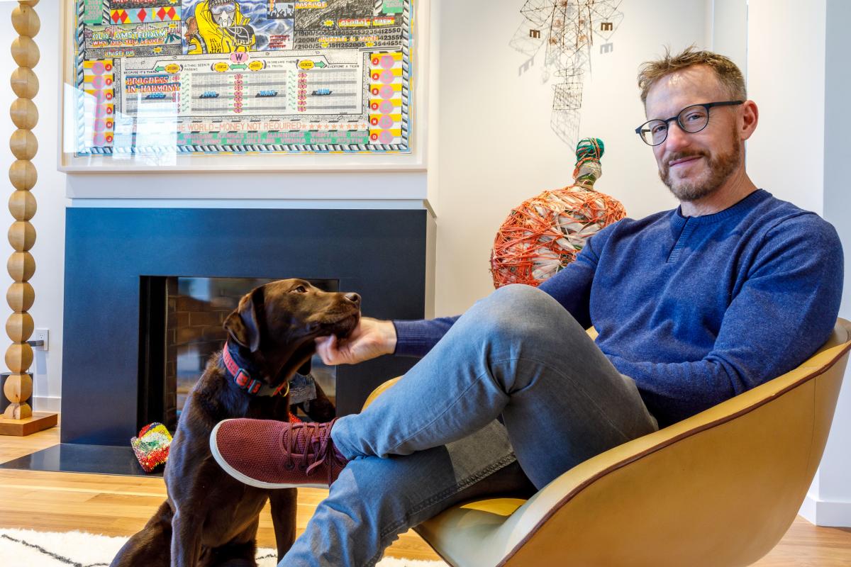 A man in a blue sweater and jeans sits in a light brown chair with his dog by his side, surrounded by works by self-taught artists.
