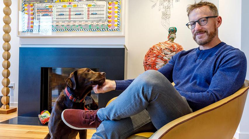 A man seated in his living room, petting his dog.