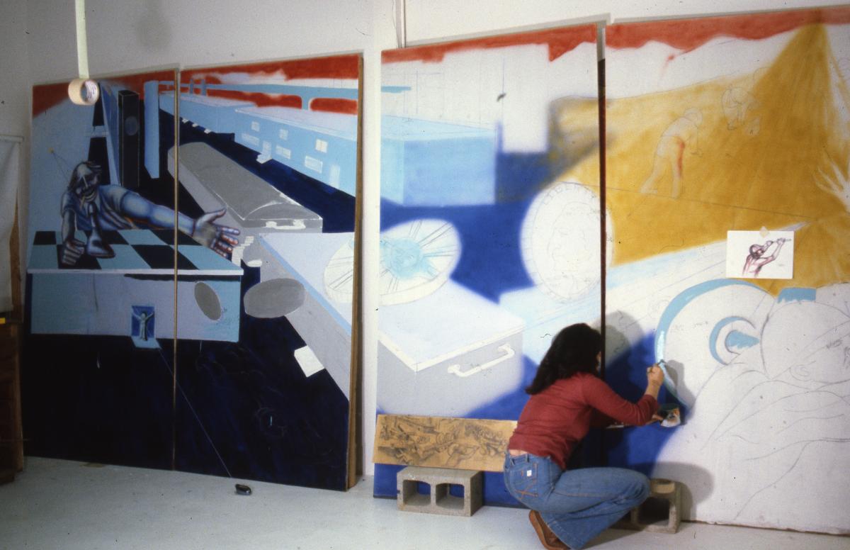 Artist Judith Baca in jeans and red shirt kneels and works on a panel of her mural, Uprising of the Mujeres