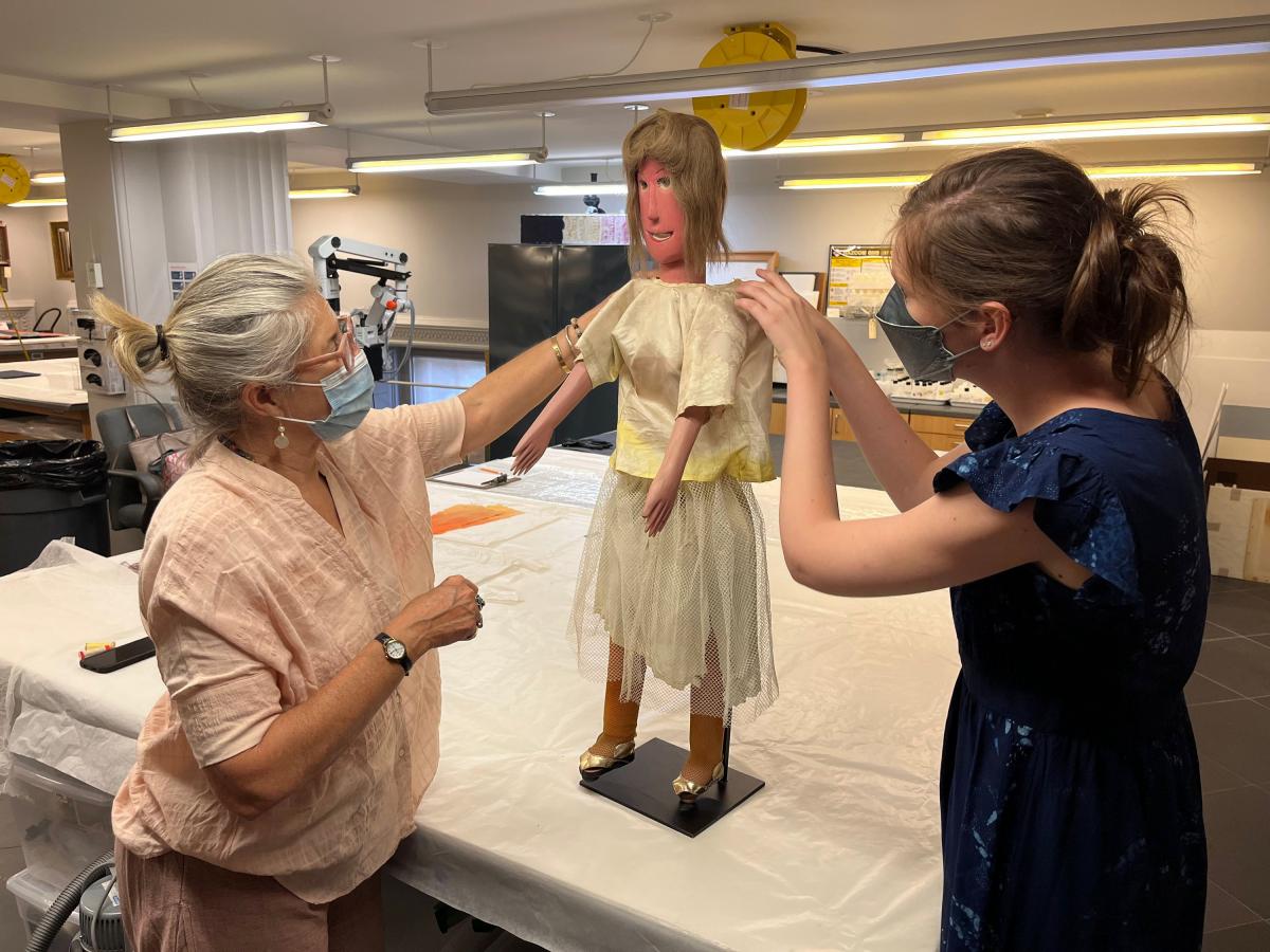 Two conservators, both wearing masks, re-dress the Gladys doll.