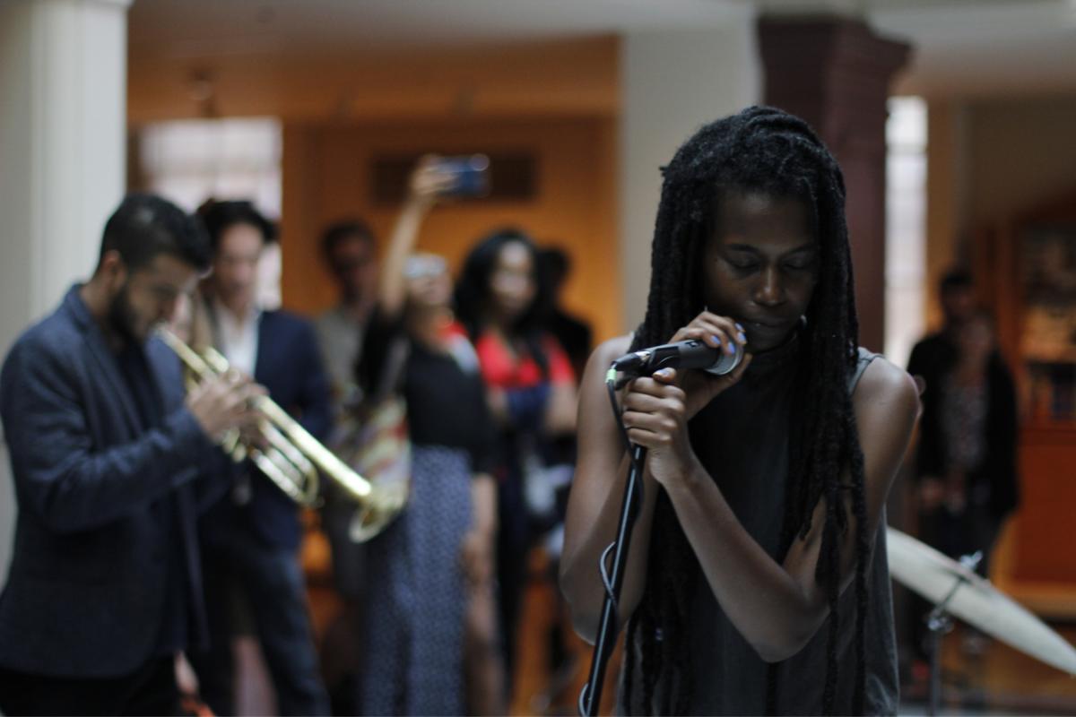 A woman singing into a microphone with a man playing trumpet behind her.