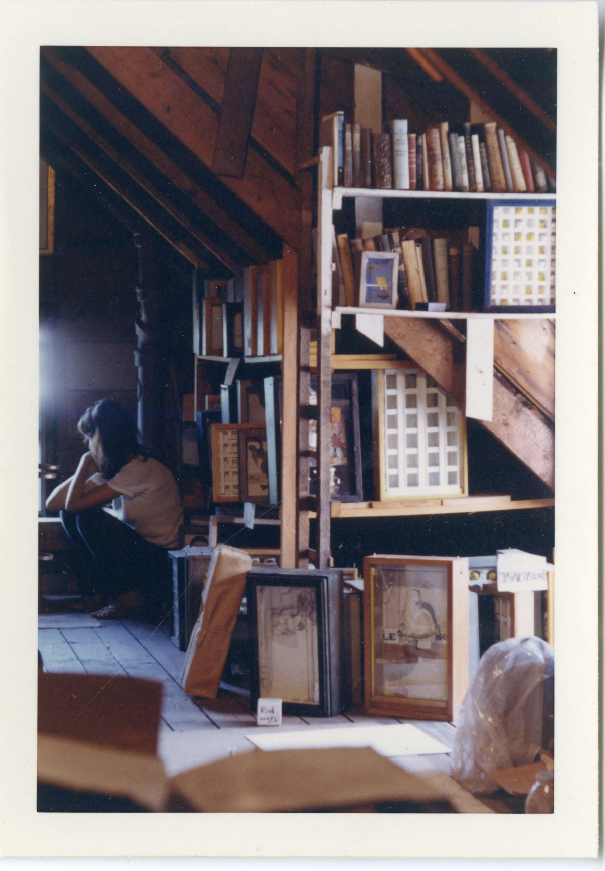 A view of an attic with filled shelves, boxes and artworks on the floor, an unidentified person sitting in the background