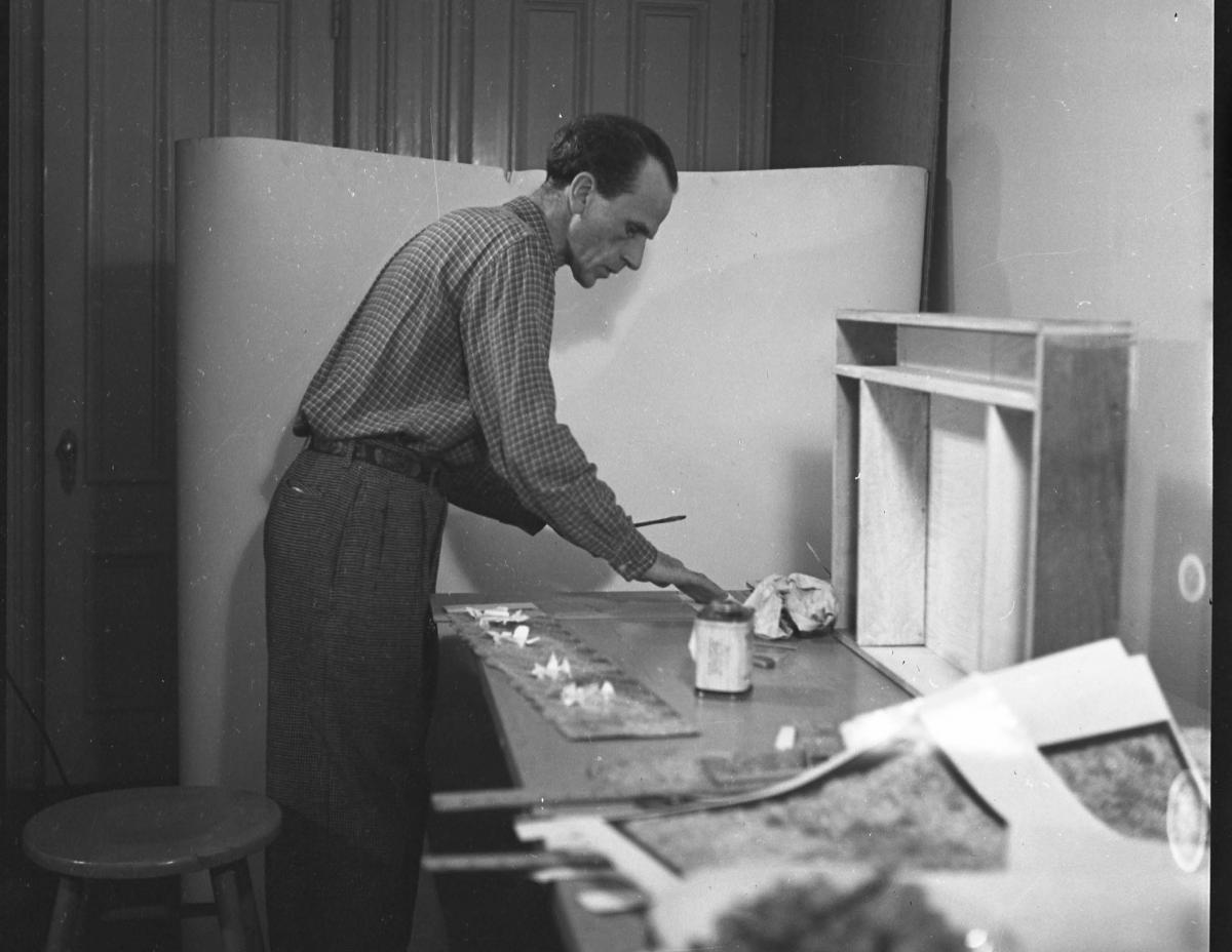 A black-and-white photo of an artist standing over his work desk with boxes and other materials
