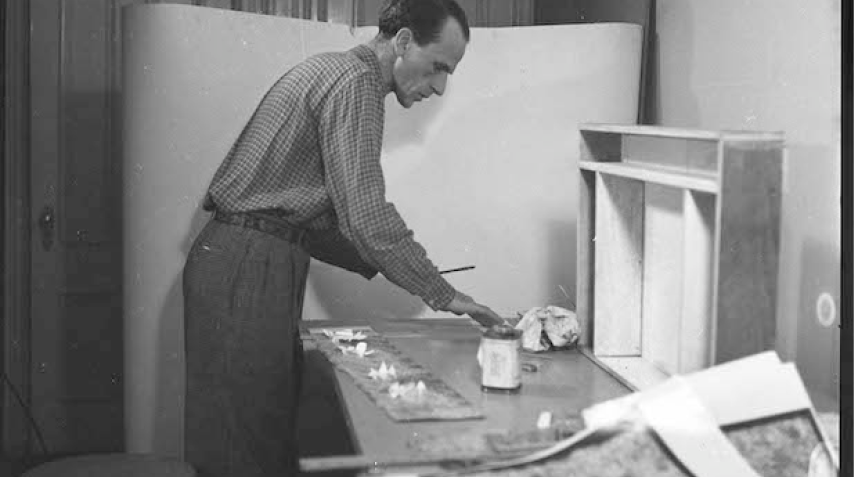 Artist Joseph Cornell standing at worktable with materials for his artworks around him