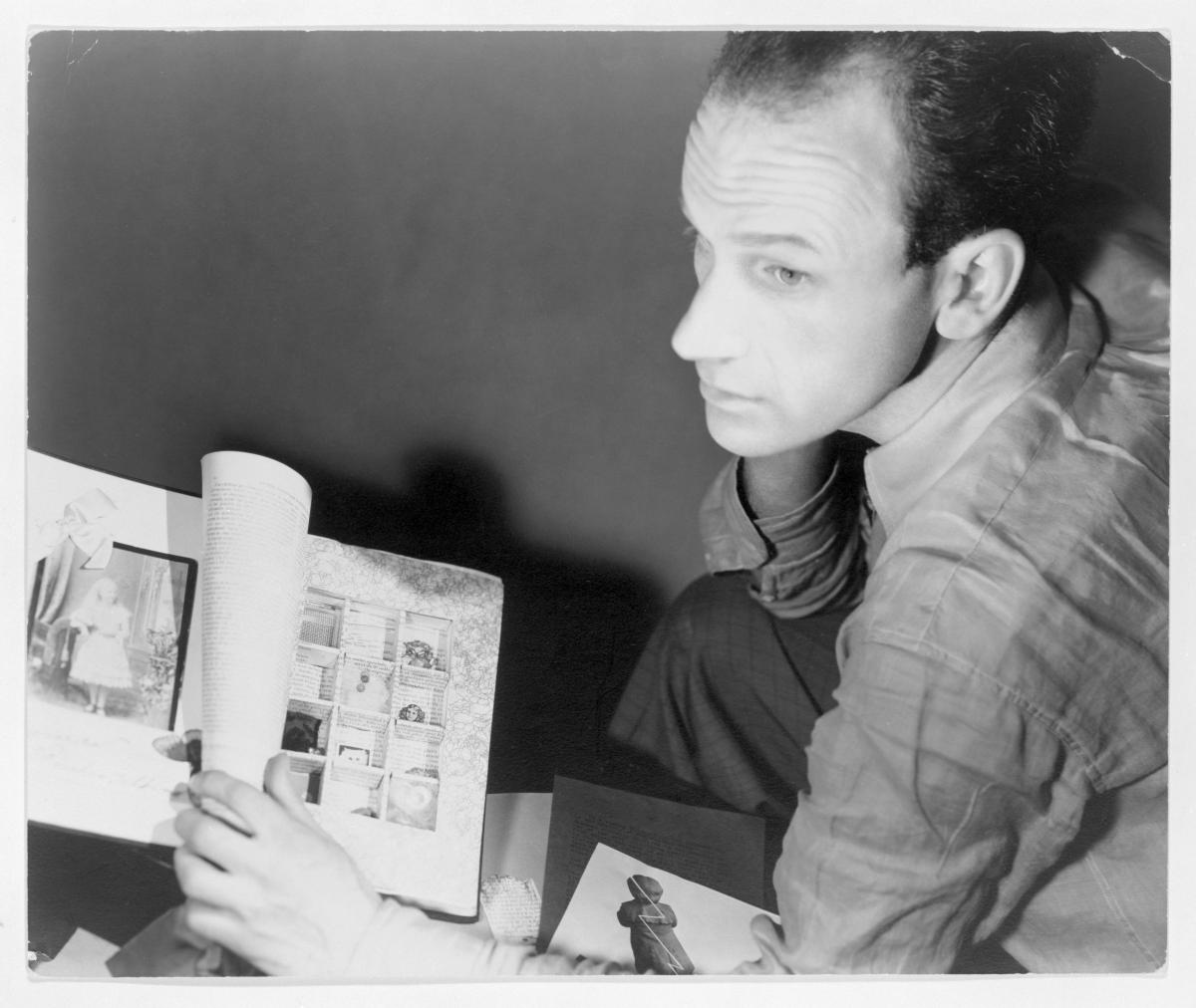 A black and white photo of a man sitting and looking away from the camera, an assemblage artwork at his side