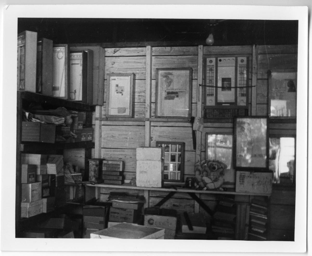 A black and white photo of artworks framed on walls, some on a table, some stacked on the floor